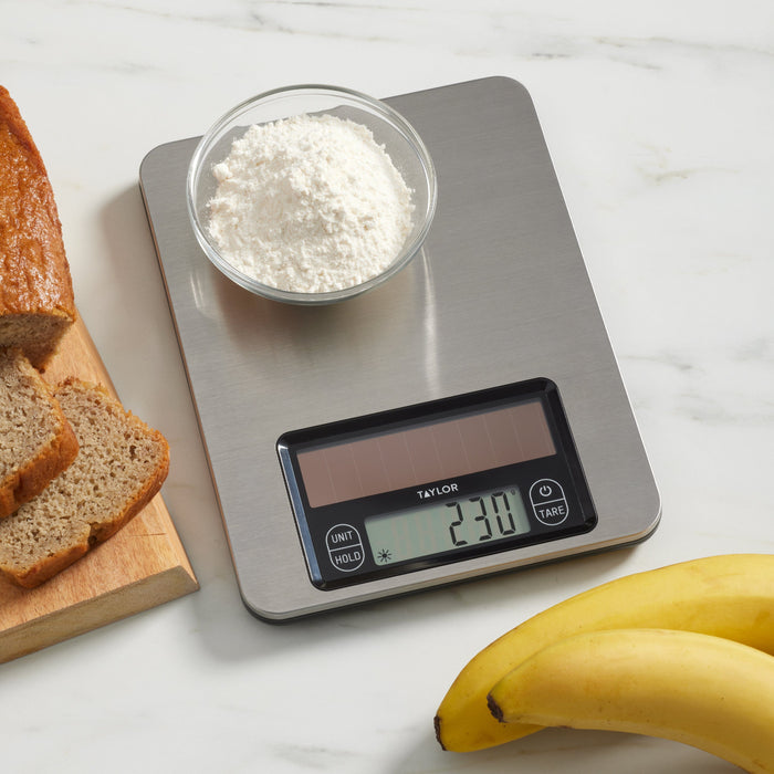 Brushed stainless steel kitchen scale with a bowl of flour on the weighing surface and bread nearby.