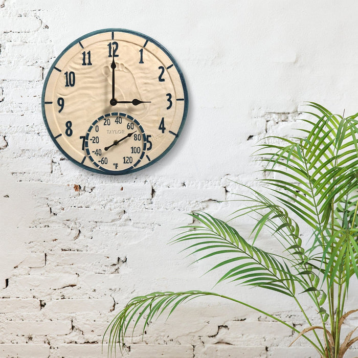 14 inch coastal clock with built-in thermometer mounted on a white brick wall beside a green plant