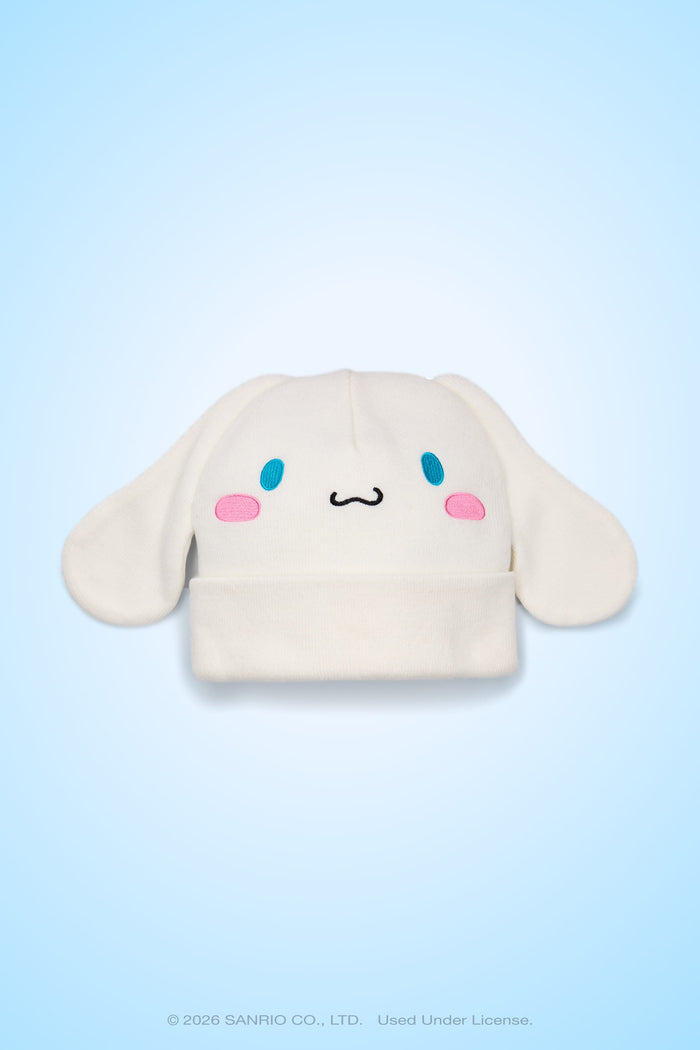 The 686 Women’s Cinnamoroll™ Beanie by 686 features a white acrylic knit design with floppy ears, blue oval eyes, pink cheeks, and a small smiling mouth that resembles the cute cartoon character Cinnamoroll, shown against a light blue background.