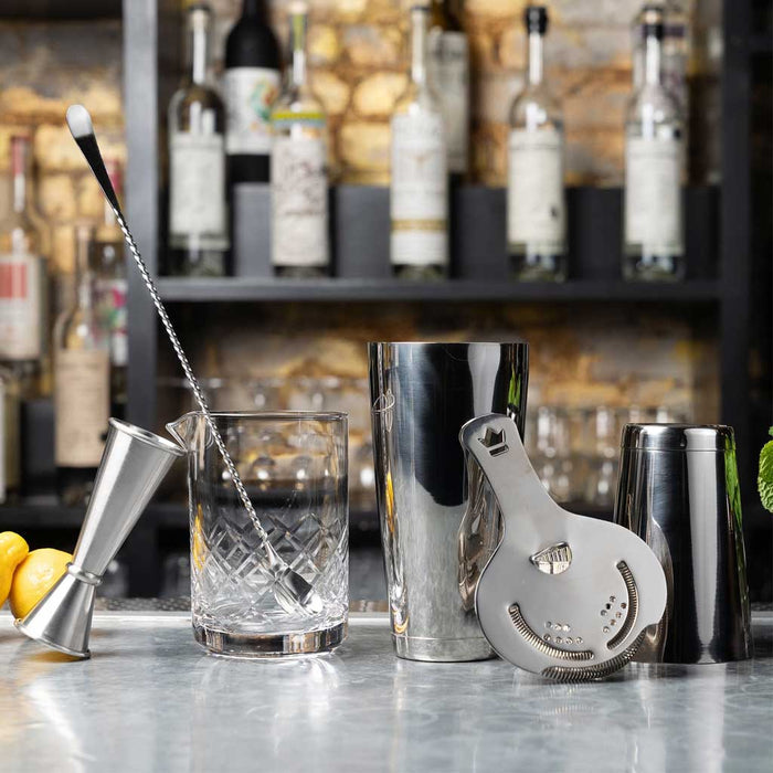 Bar tools including a jigger, cocktail shaker, and strainer on a bar counter with bottles in the background.
