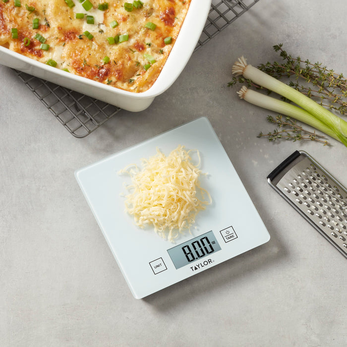 Compact Taylor kitchen scale with a white weighing platform and digital display reading 0.0 oz beside a casserole dish