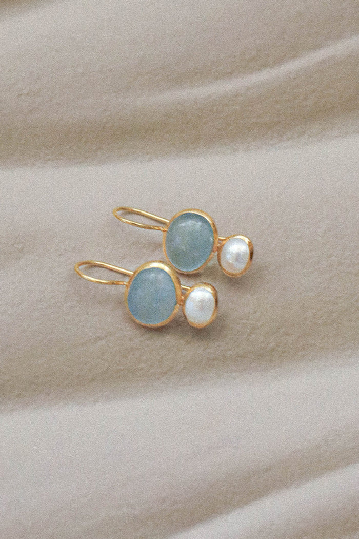 A pair of gold-tone drop earrings featuring oval light blue gemstones and small white pearl-like beads, displayed on a soft beige fabric background.
