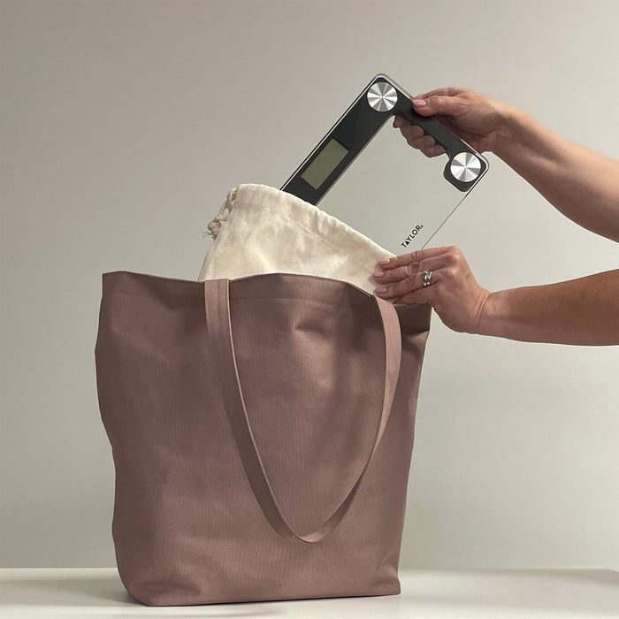 Person weighing a tan tote bag using a digital scale on a table
