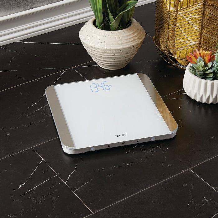 Glass digital bathroom scale on dark marble floor beside a speckled white pot with a green plant and a gold decorative lamp