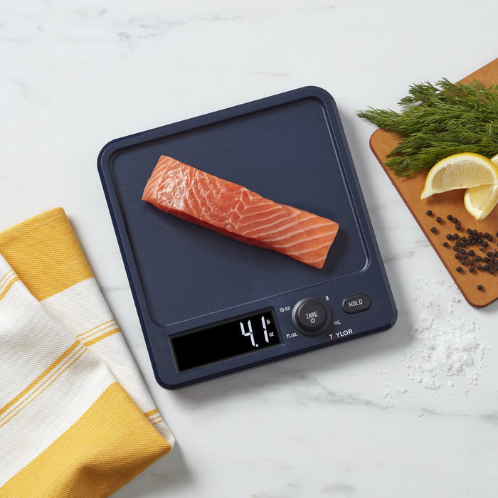 Salmon fillet resting on a dark navy antimicrobial kitchen scale with rotating knob and a yellow towel nearby