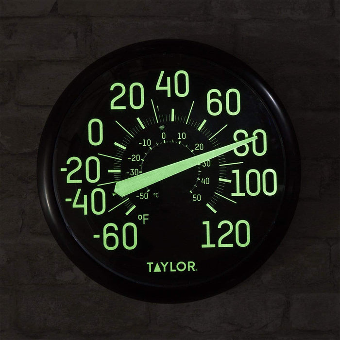 Large glow in the dark Taylor outdoor thermometer with bright green numerals on a black dial against a brick wall.