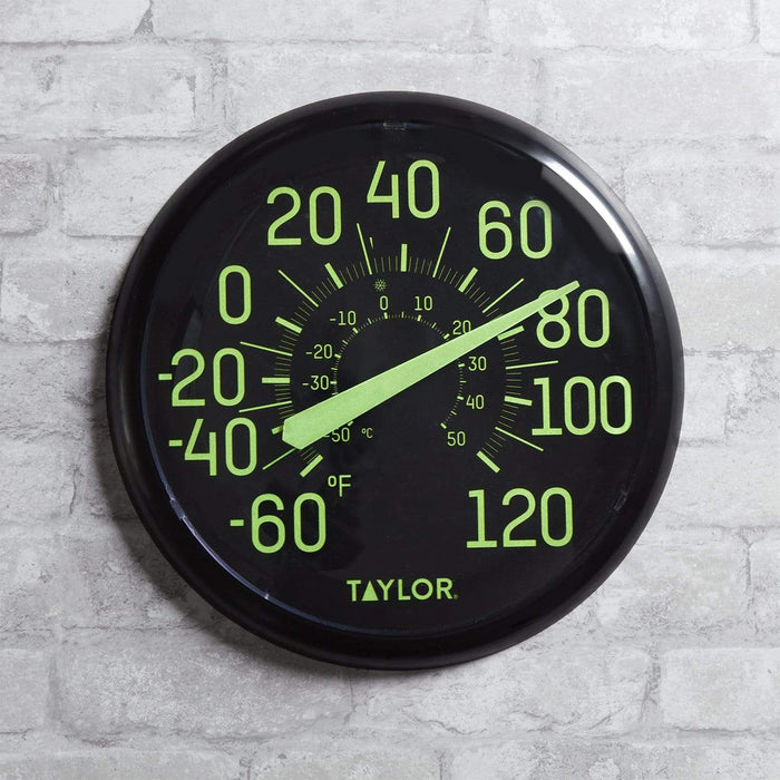Round black Taylor glow in the dark outdoor thermometer with neon green numbers mounted on a light brick wall.