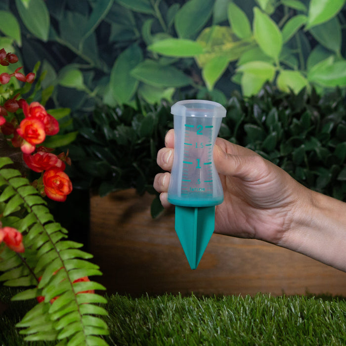 Hand holding a 2 inch silicone sprinkler gauge with green markings in a garden background.