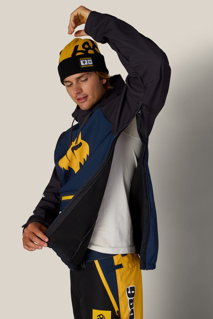 A person wearing a yellow and black beanie and a 686 Men’s Fox Racing Waterproof Hoody, demonstrating the jacket’s side-zip ventilation.
