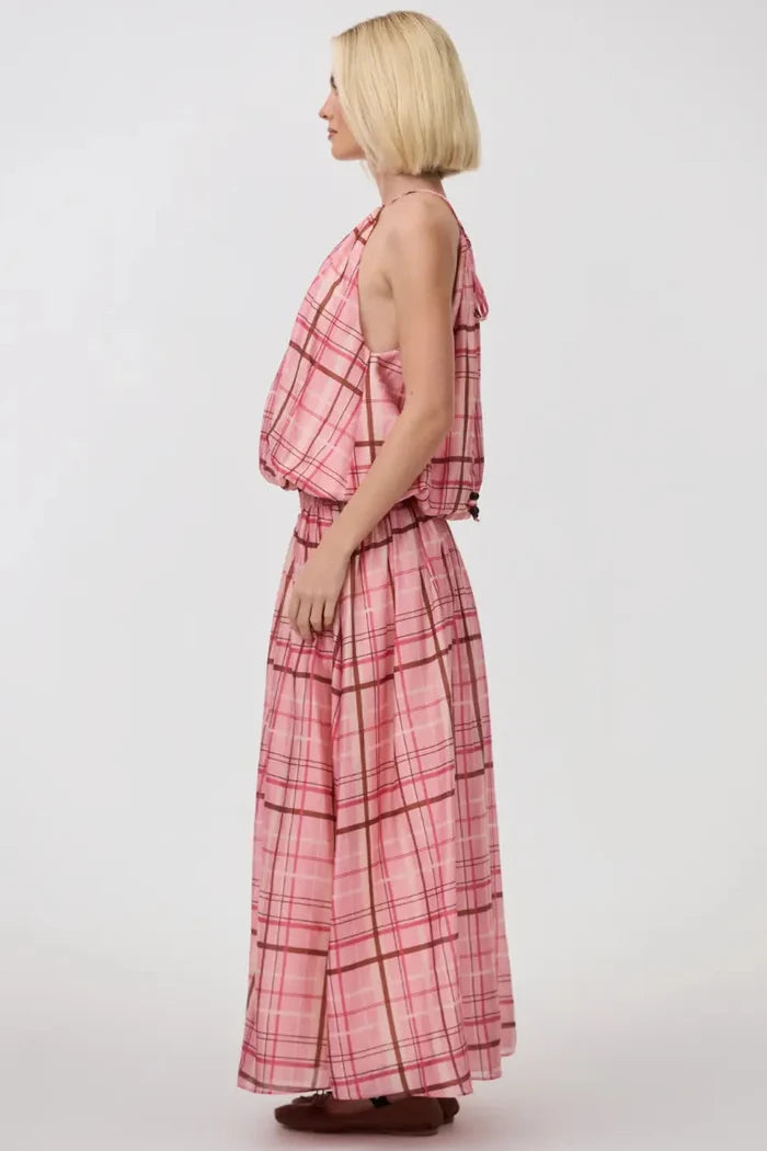A woman with a short blonde bob is shown from the side profile wearing a pink and red plaid halter-neck maxi dress with a matching belt and skirt, standing against a plain light gray studio background.