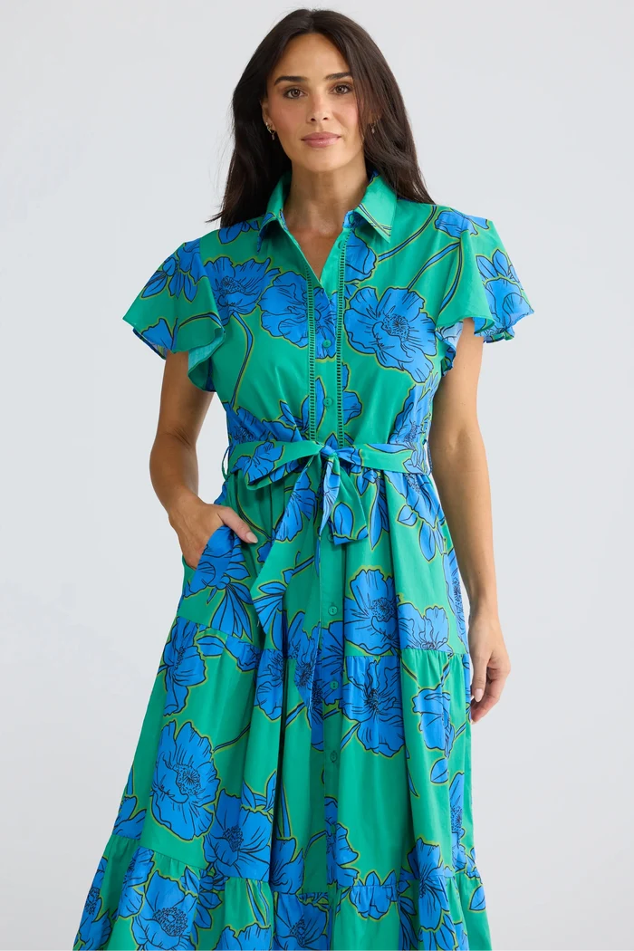 A woman modeling a green and blue floral print midi dress with a tie waist and short flutter sleeves, set against a plain light gray studio background.