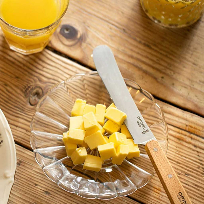 Butter cubes on a glass plate with an Opinel brunch knife, next to a glass of orange juice on a wooden surface.