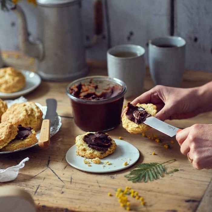 Person spreading chocolate on a scone with an Opinel brunch knife with a wooden table setting and tea cups in the background.