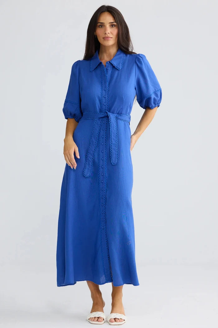 A woman modeling a vibrant blue midi shirt dress with puff sleeves, a button-up front, and a matching tie belt, set against a plain light gray studio background.