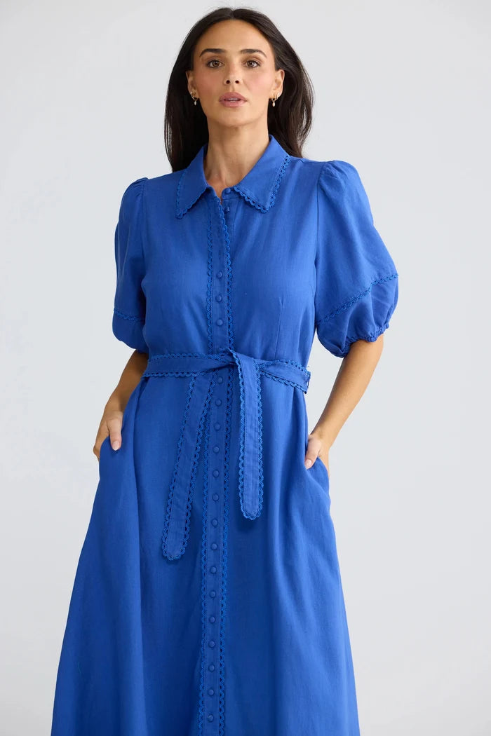 A woman modeling a bright blue midi dress with puff sleeves, a pointed collar, and a matching waist tie, set against a plain light gray studio background.