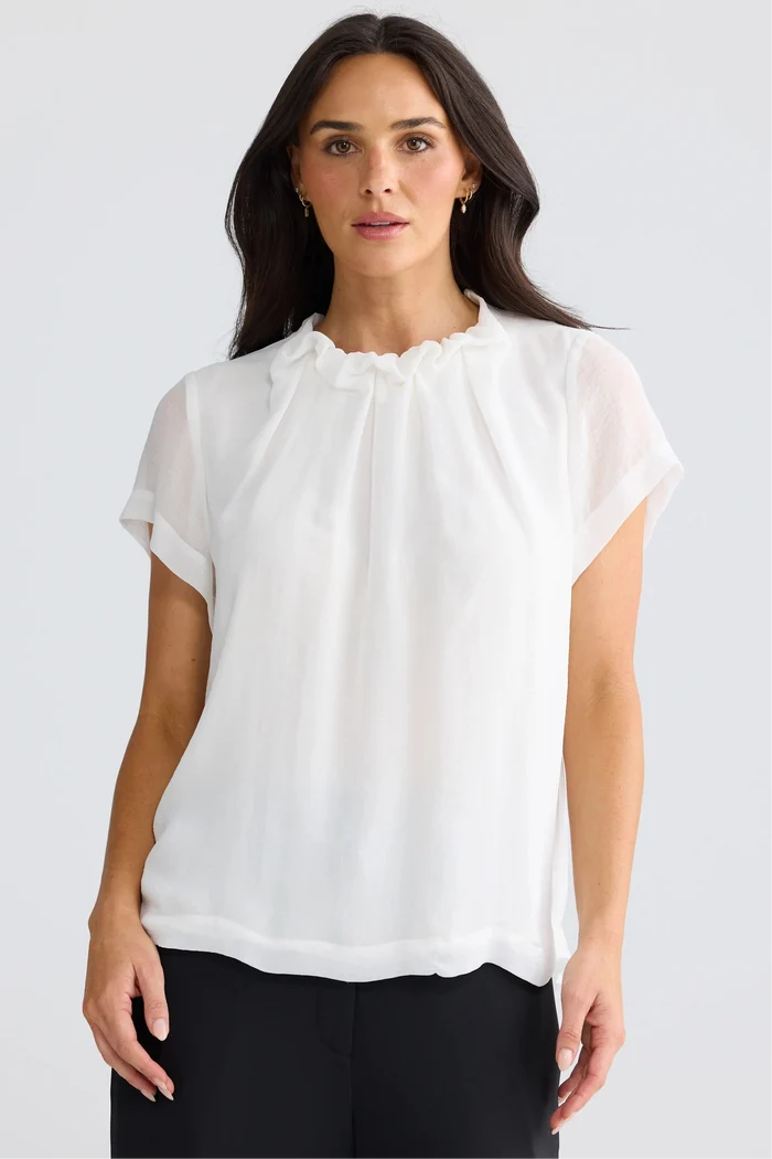 A woman wearing a white short-sleeve blouse with a ruffled high neckline, paired with black pants, photographed against a light gray studio background.