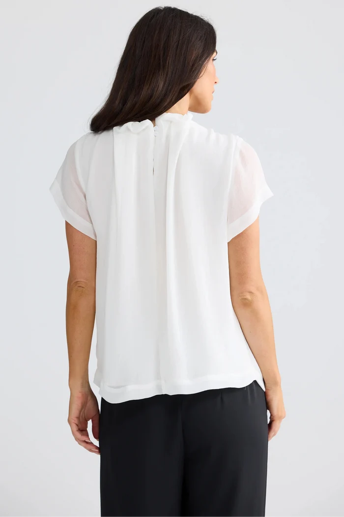 A woman viewed from the back wearing a white short-sleeve blouse with a gathered, pleated back detail and black pants, set against a plain light gray studio background.