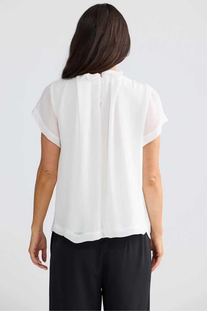 A woman viewed from the back wearing a white short-sleeve blouse with a gathered, pleated back and black pants, set against a plain light gray studio background.