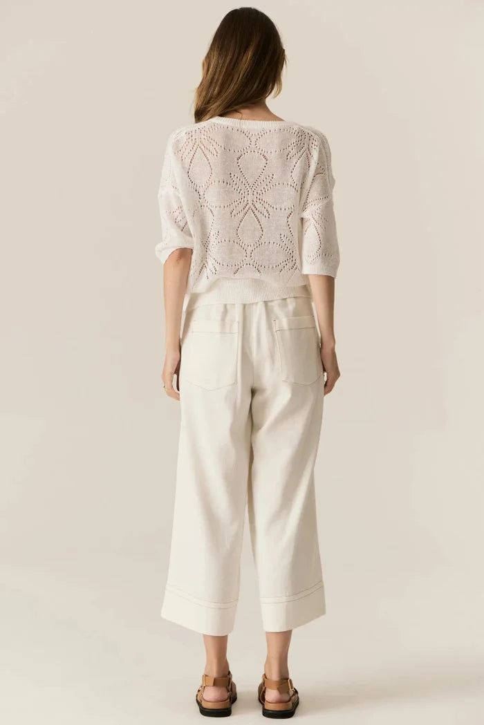 Woman wearing a light pink lace top and white pants against a beige background