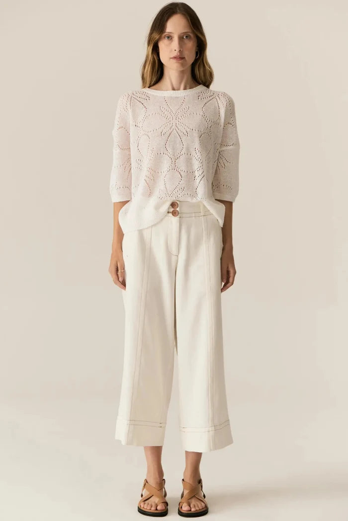 Woman wearing a light pink knitted top and white pants on a plain background