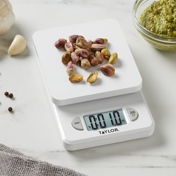 White compact kitchen scale on a marble surface with pistachios on the weighing platform and a bowl of pesto nearby