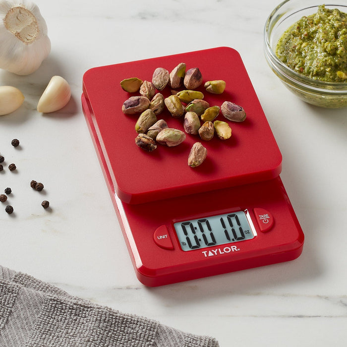 Red Taylor compact digital kitchen scale topped with pistachios on a white marble counter with a pesto bowl nearby
