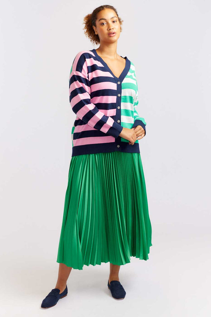 A woman standing and posing in a studio setting, wearing a pink and green striped sweater and a green pleated skirt, paired with blue shoes.