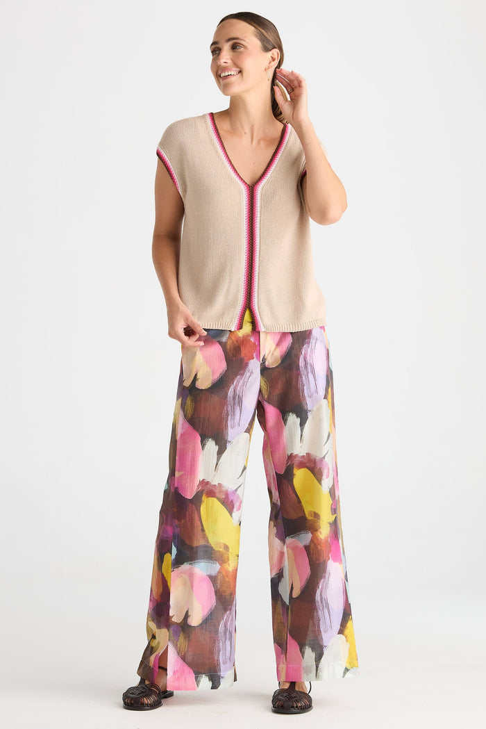 A woman modeling a beige sleeveless knit top with pink trim and matching wide-leg pants featuring a colorful abstract floral print in pink, yellow, purple, and brown, set against a plain white studio background.