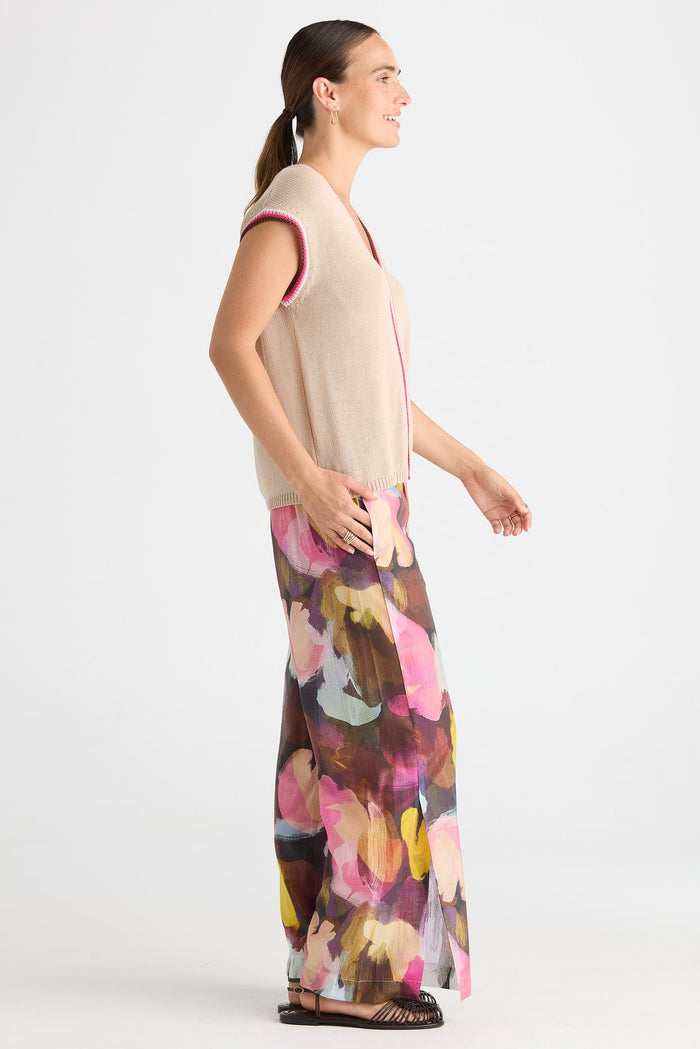 A woman shown in profile wearing a sleeveless beige knit top with pink trim and wide-leg floral print pants in pink, yellow, brown, and white, photographed against a light gray studio background.