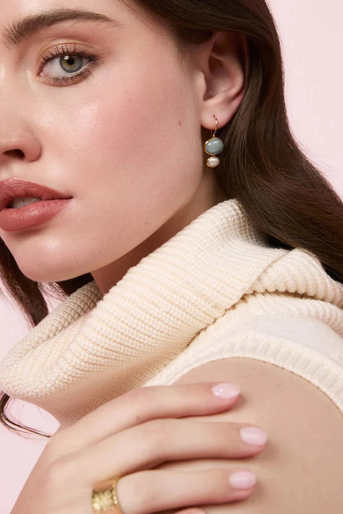 A close-up of a woman wearing a cream ribbed knit cowl neck sweater vest, a gold ring, and a light blue and white beaded drop earring, set against a solid pale pink background.