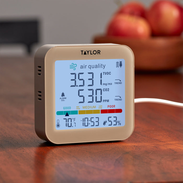 Taylor digital air quality monitor with blue display showing air quality metrics on a wooden table with blurred apples in the background