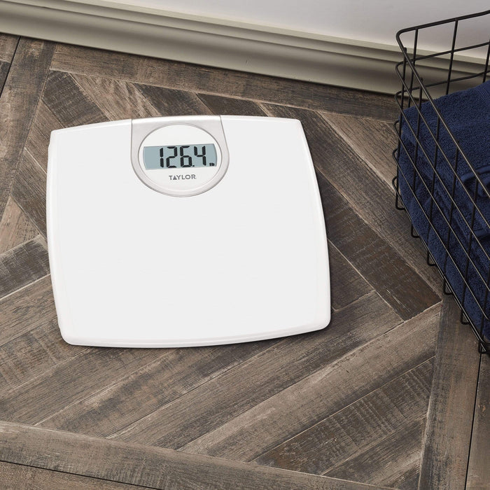 White digital bathroom scale on wood floor beside a wire laundry basket displaying 126.4 lb