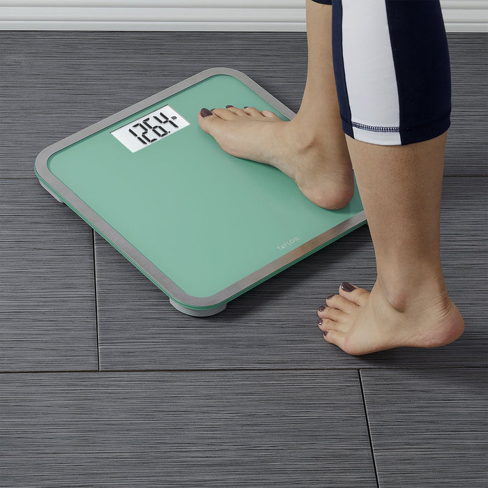 Foot on a seafoam green digital glass scale on a wooden floor; person wearing navy and white athletic leggings
