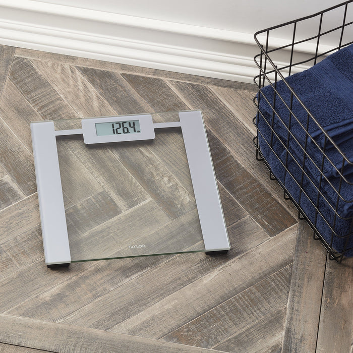 Angled view of a digital glass scale on a wood floor with a metal basket and navy towel nearby