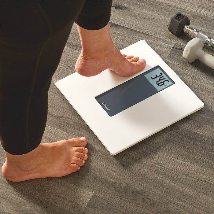 Bare feet on a white digital scale on a wood floor with a dumbbell nearby.
