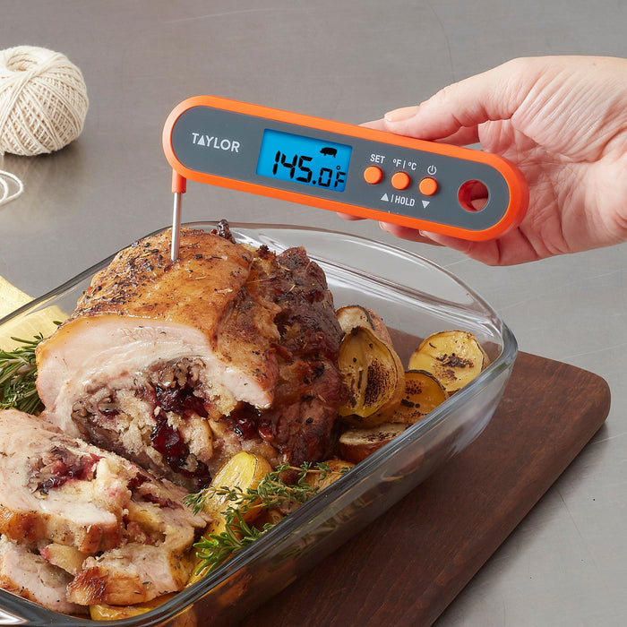 Hand holding orange digital thermometer over a roasted chicken with potatoes and rosemary in a glass dish.