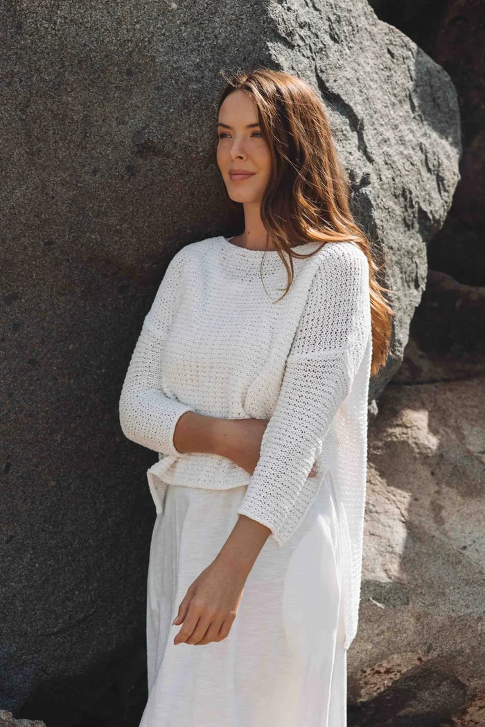A woman standing by a large rock, wearing a white Driftaway Pullover with a high low hem and boat neck, with rolled-up sleeves.