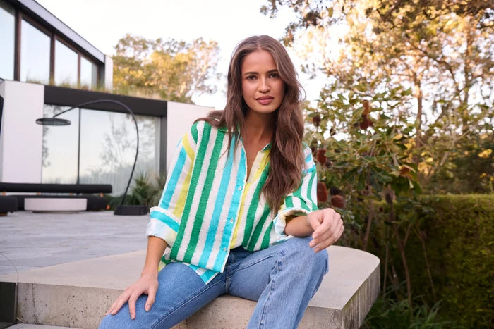 Woman sitting outdoors wearing a colorful striped shirt and jeans.
