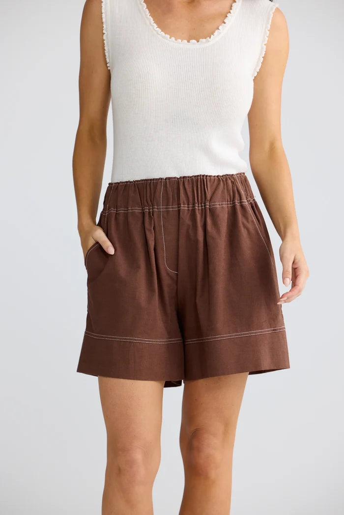A model wearing brown linen-look shorts with contrast stitching and a white sleeveless top, shown from the chest down against a light gray studio background.