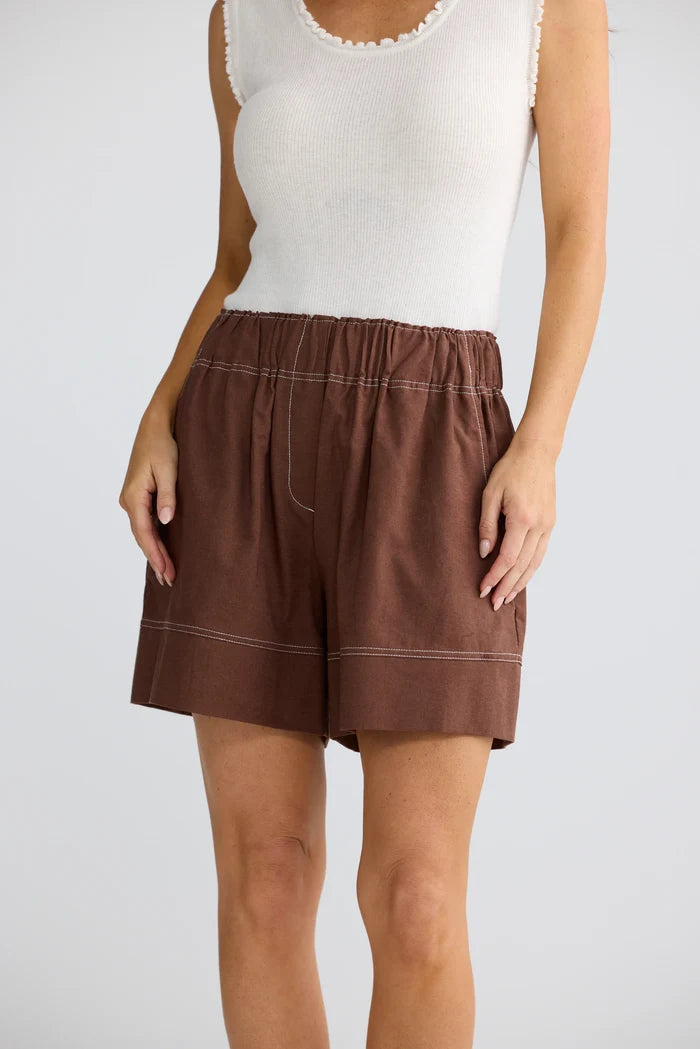 A model wearing brown linen shorts with white contrast stitching and a white sleeveless top, shown from the chest down against a light gray background.
