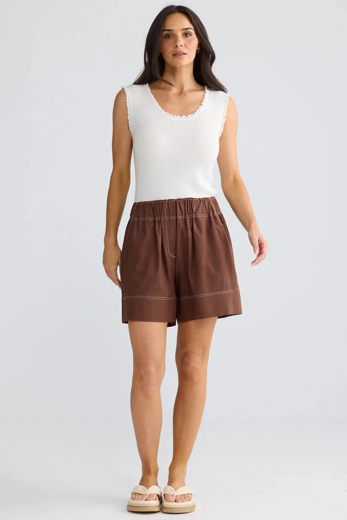 A woman modeling a sleeveless white knit top and brown linen shorts against a light gray studio background.