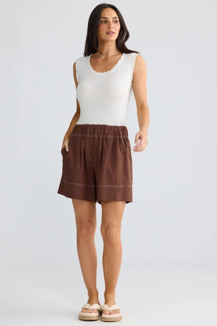 A woman modeling a sleeveless white top and brown shorts against a light gray studio background.
