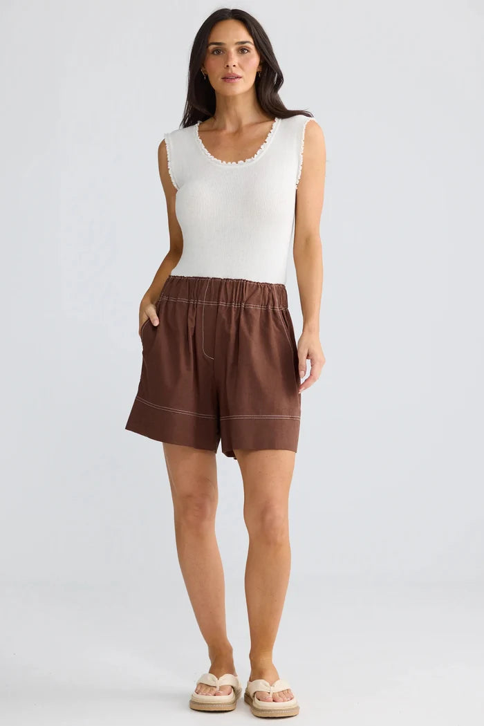 A woman modeling a white sleeveless knit top and brown linen shorts against a light gray studio background.