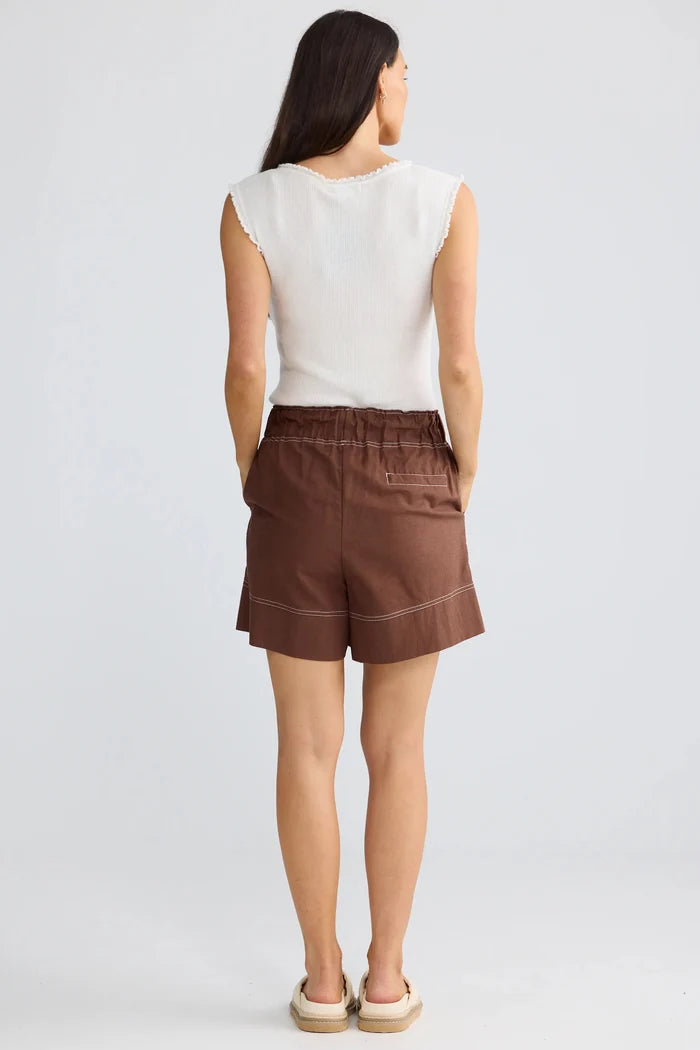 A woman is shown from behind wearing brown shorts and a white sleeveless top, standing against a light gray studio background.