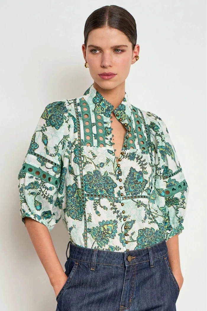 A woman modeling a short-sleeved button-up blouse with a green and teal floral paisley print on a white background, paired with dark blue jeans against a light gray studio background.