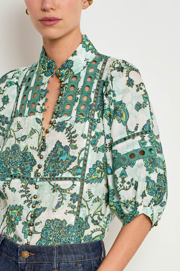 A close-up of a woman wearing a high-neck blouse with a white base and a teal and green botanical print, featuring decorative cutouts and button details, paired with dark blue jeans.