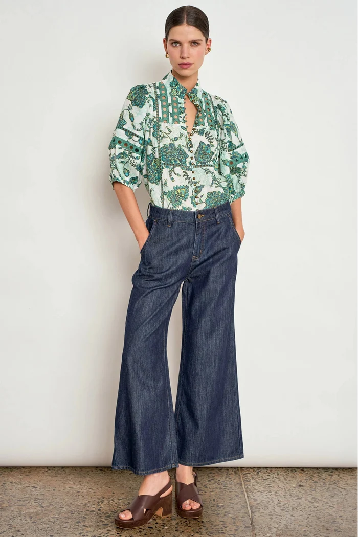 A model stands against a white wall wearing a green and white botanical print blouse with a keyhole neckline, dark blue wide-leg denim pants, and brown heeled sandals.
