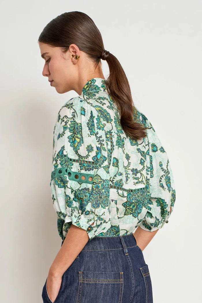 A woman shown from the back wearing a light green and teal patterned blouse with dark blue jeans, set against a plain light gray studio background.