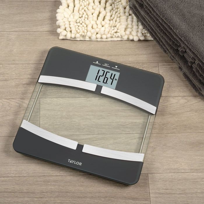 Top-down view of a glass body composition scale on a wooden floor with a white shag rug and a dark gray towel; display reads 126.4 lb