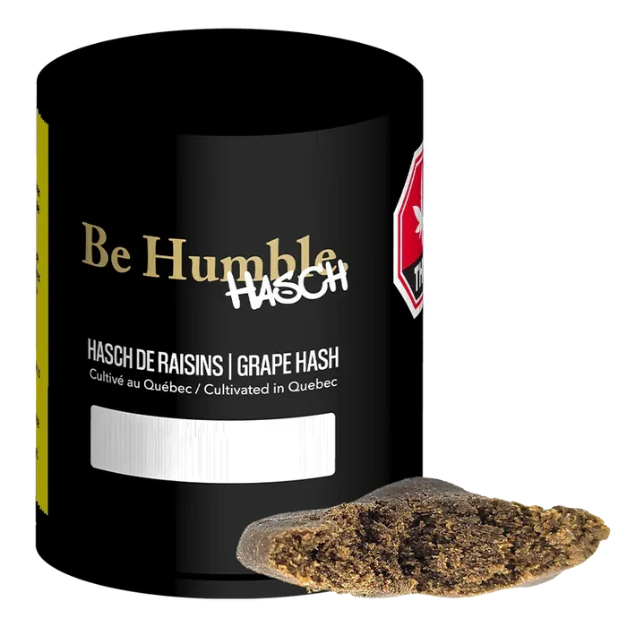Angled close-up of a black jar labeled "Grape Hash" by Be Humble with a broken brown hash piece resting in front.